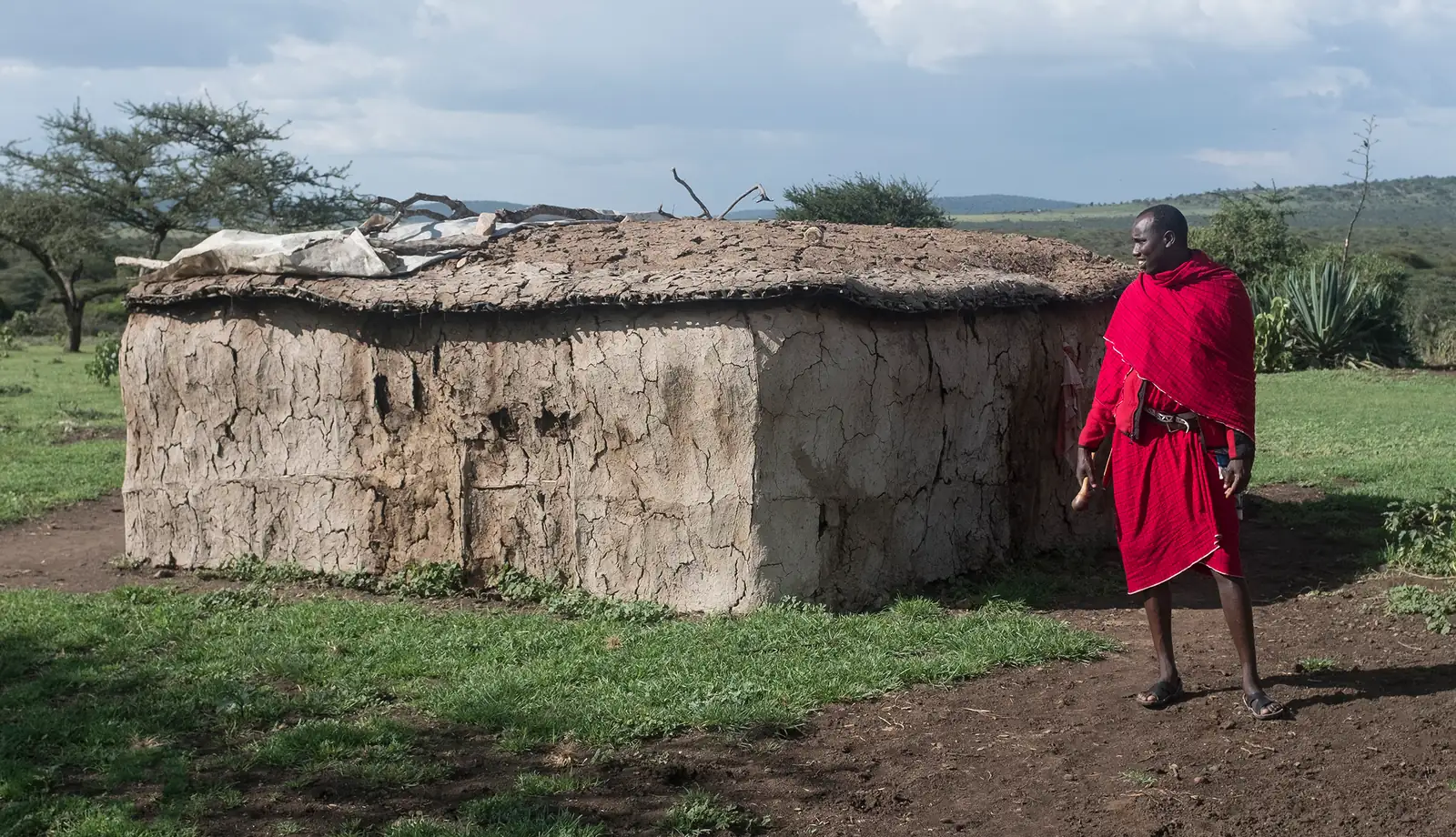 A Maasai man in a red shuka beside a mud-walled house in a boma