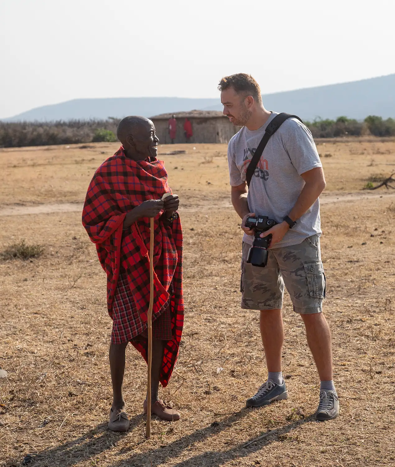 A Maasai elder in a red checked shuka talking with a guest holding a camera
