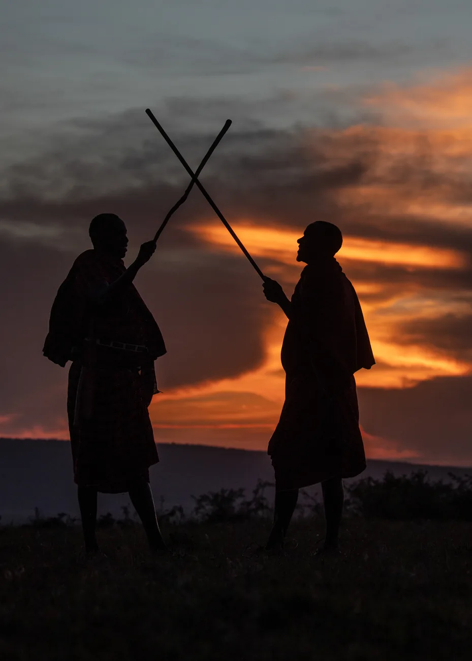 Two Maasai men crossing their sticks, silhouetted against a fiery sunset