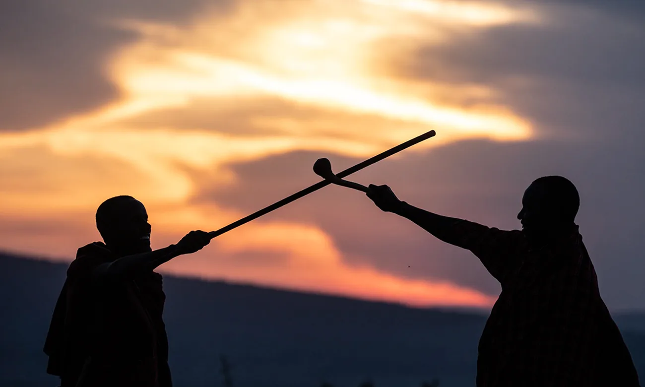 Two Maasai men silhouetted against an orange sunset, one holding out a long stick