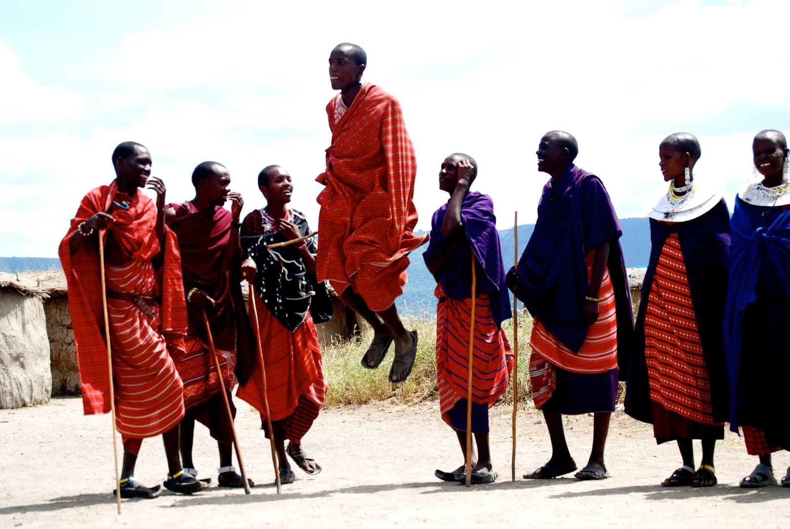 Maasai men and women in red and purple shukas, laughing as one man leaps high in the jumping dance