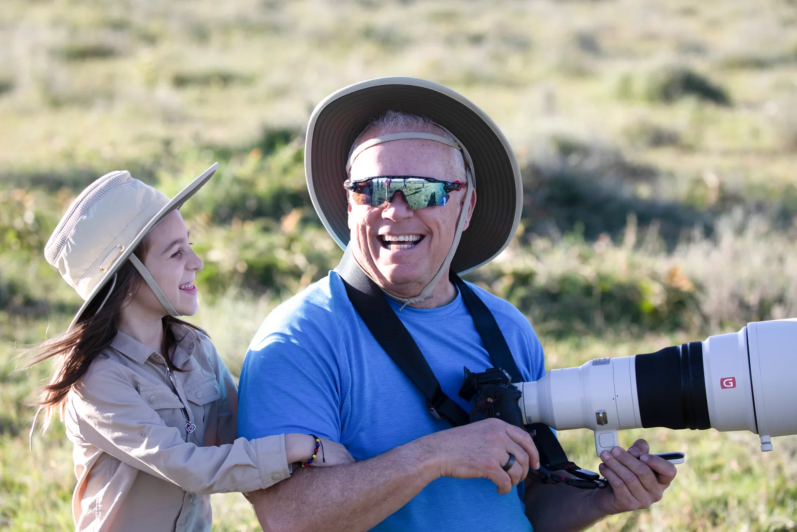 A grandfather with a long telephoto lens laughing with his granddaughter on safari