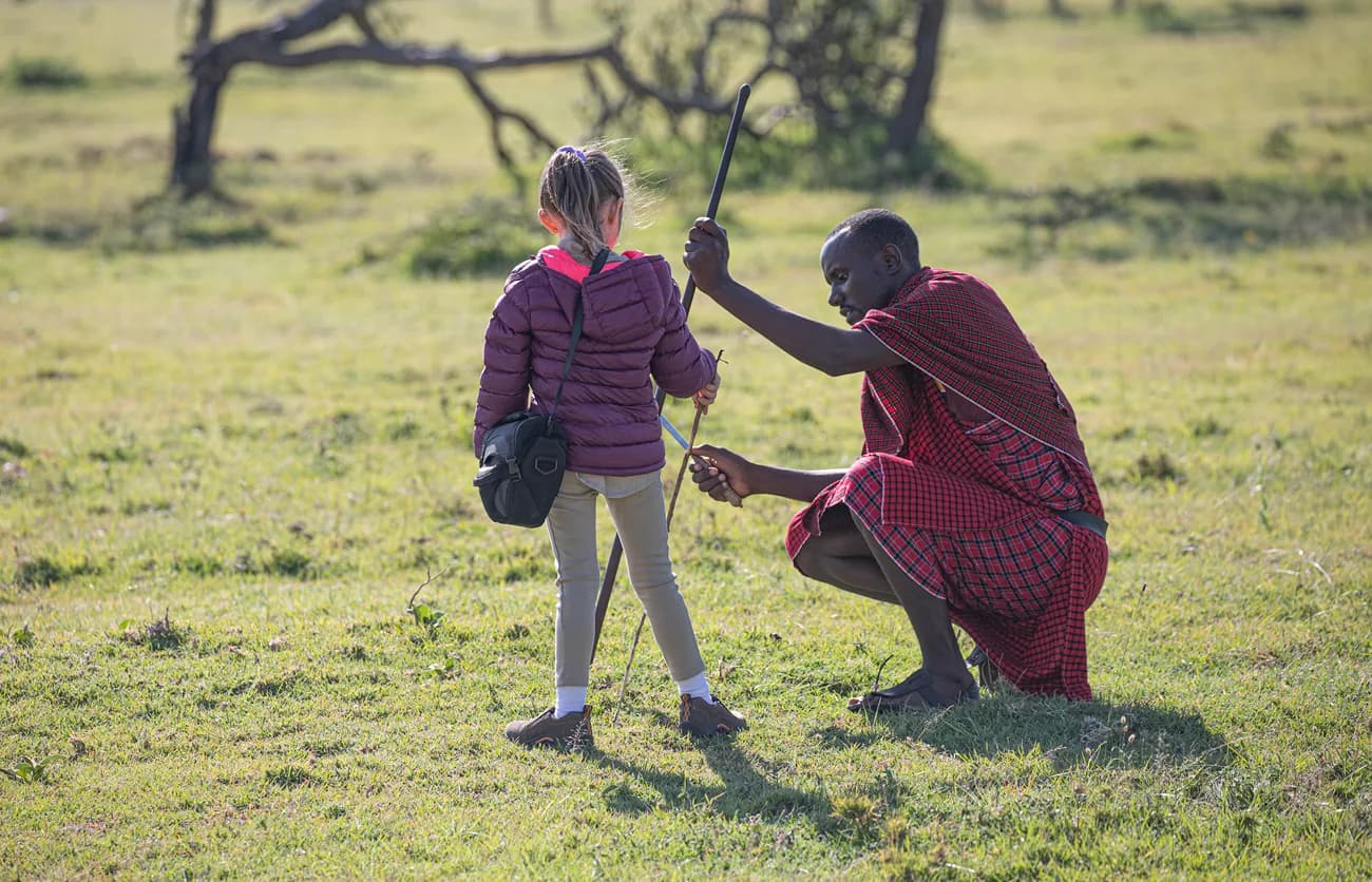 A Maasai man crouching to show a young girl how to hold a spear on open grassland