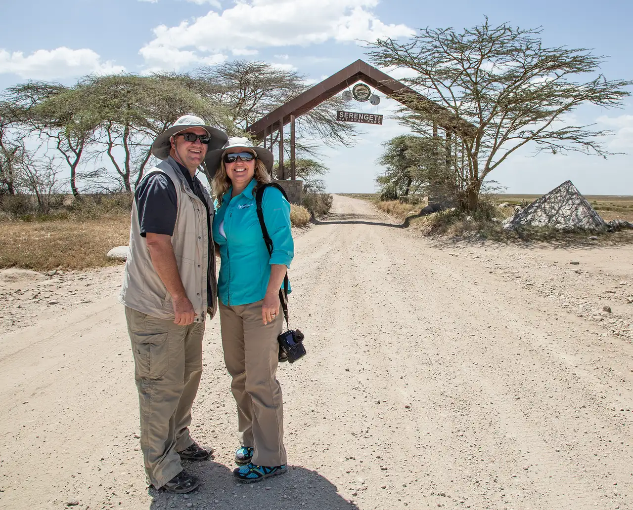 A smiling couple in safari clothes on the dirt road in front of a Serengeti park gate