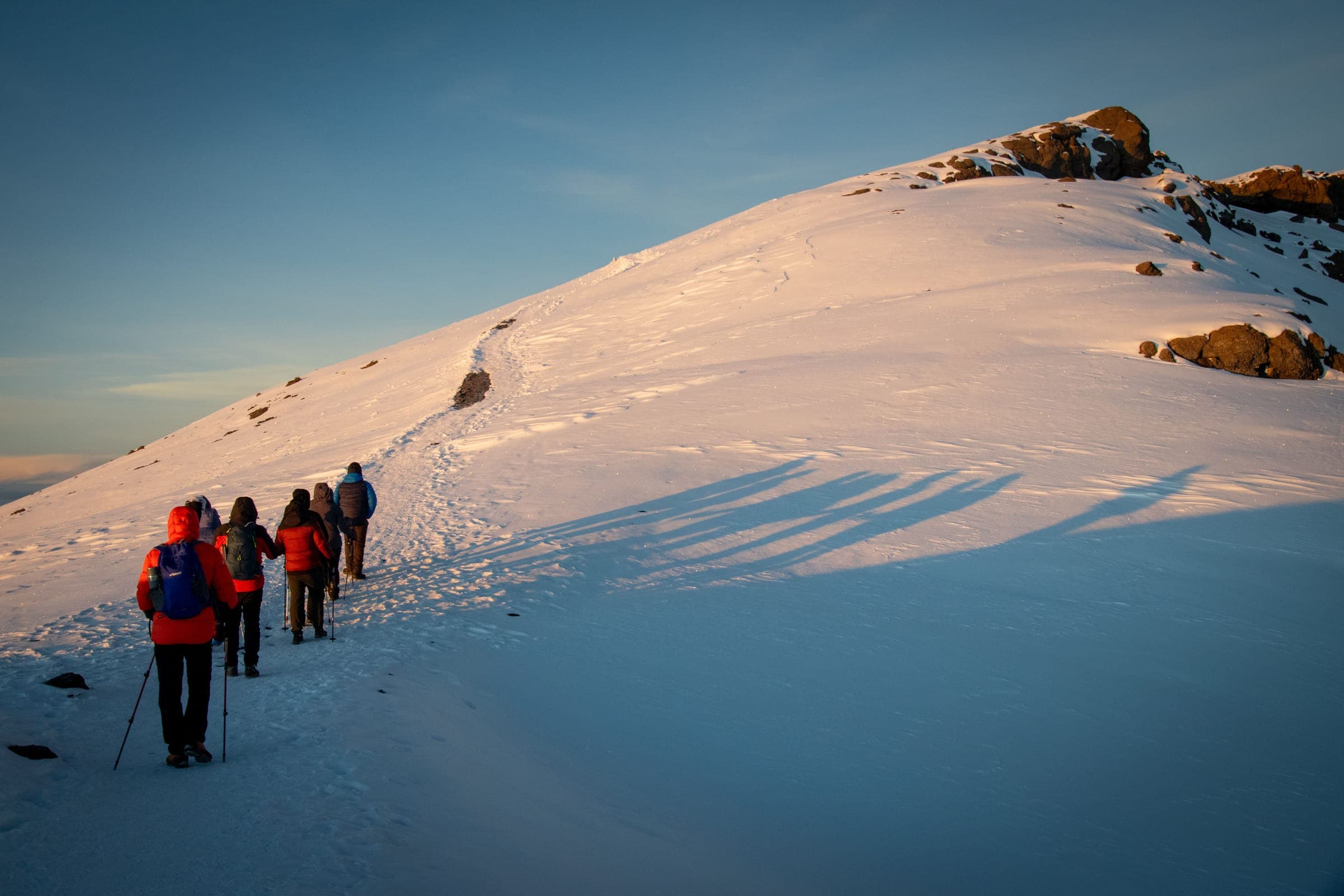 Climbers in down jackets walking up a snow slope towards a rocky summit at dawn
