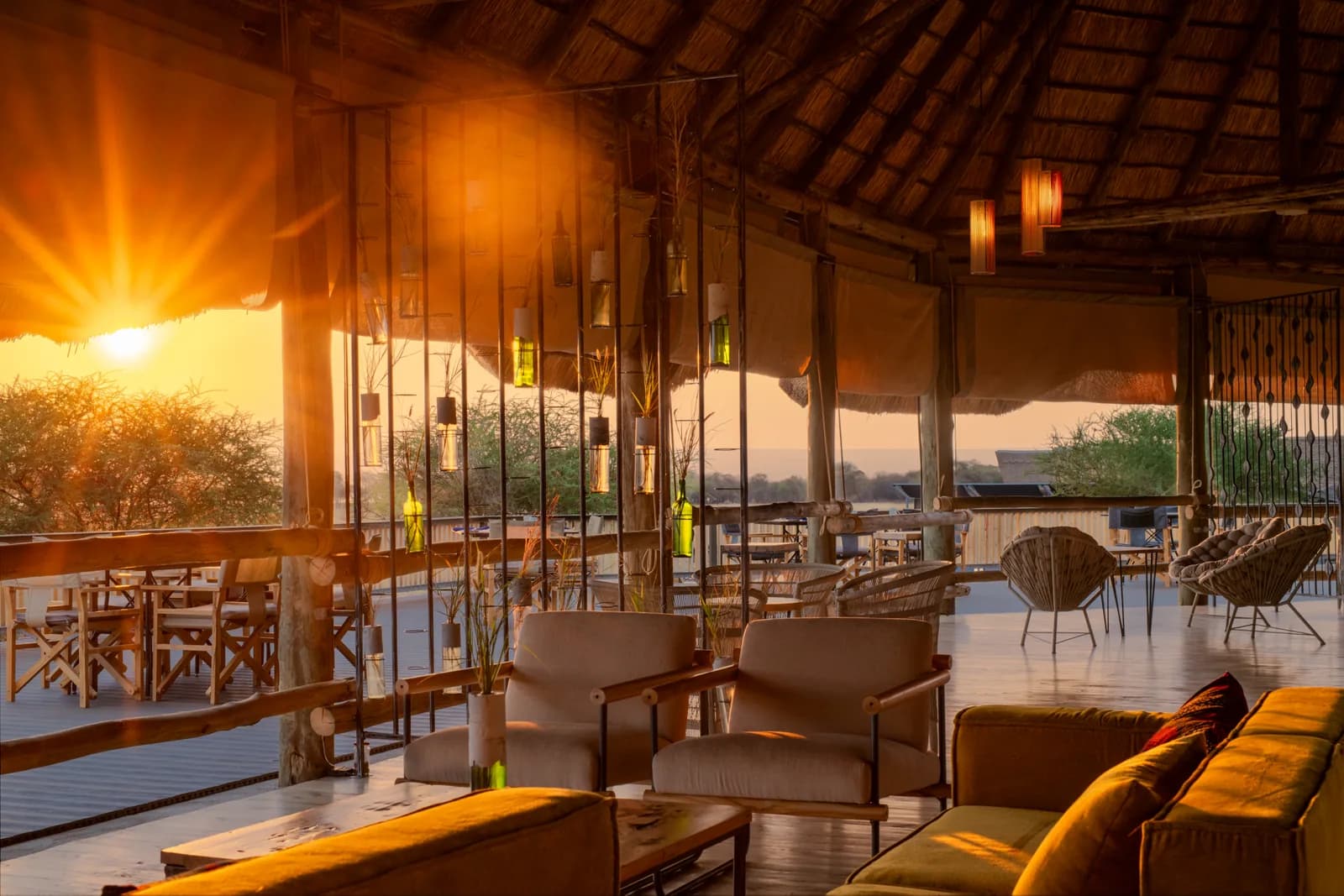 Lounge with armchairs and sofas under a high thatched roof, the setting sun behind