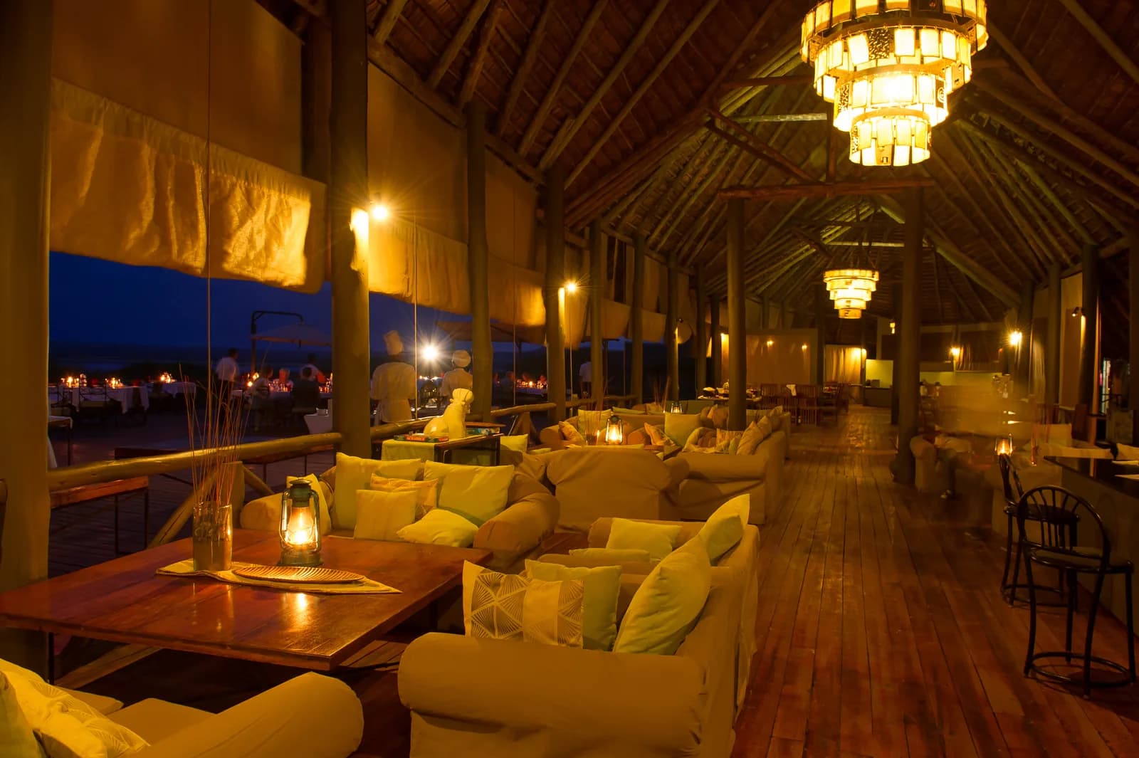 Lounge with cushioned sofas and lanterns under a high thatched roof at dusk