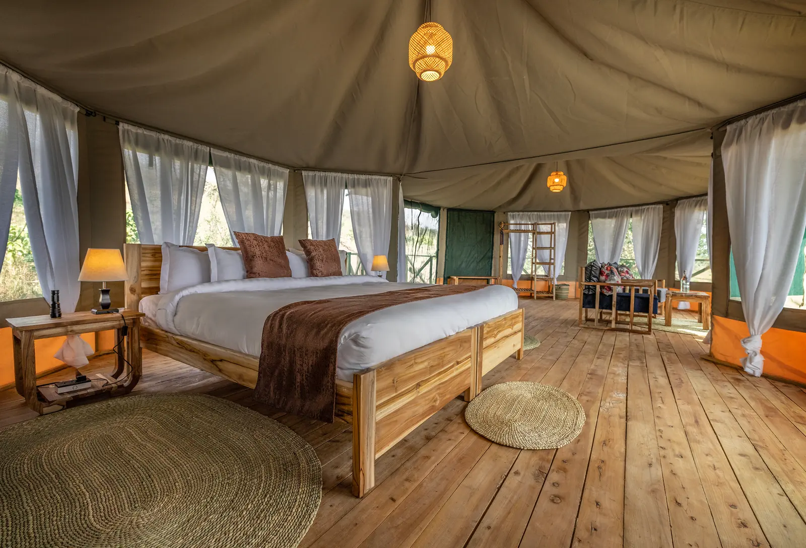 Inside a tent: a king-size bed on a wooden floor, with sheer curtains and a sitting area beyond