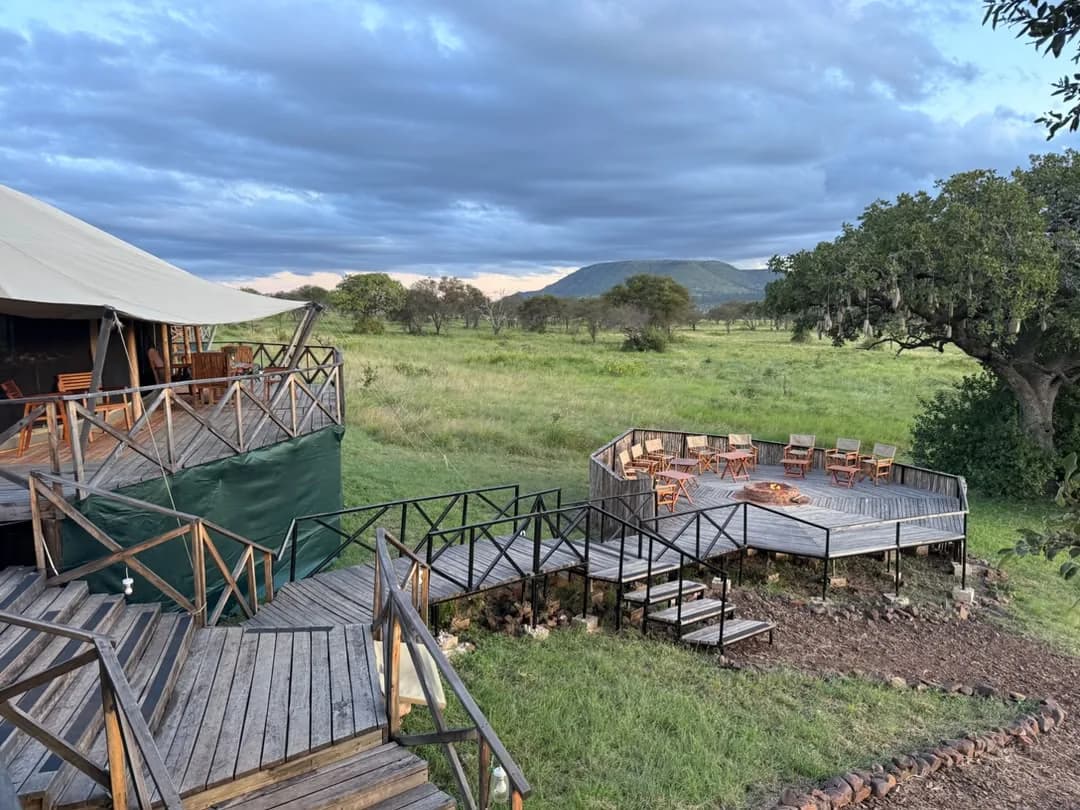 Wooden walkways from the main tent to a deck of chairs, looking over green plains to a distant hill