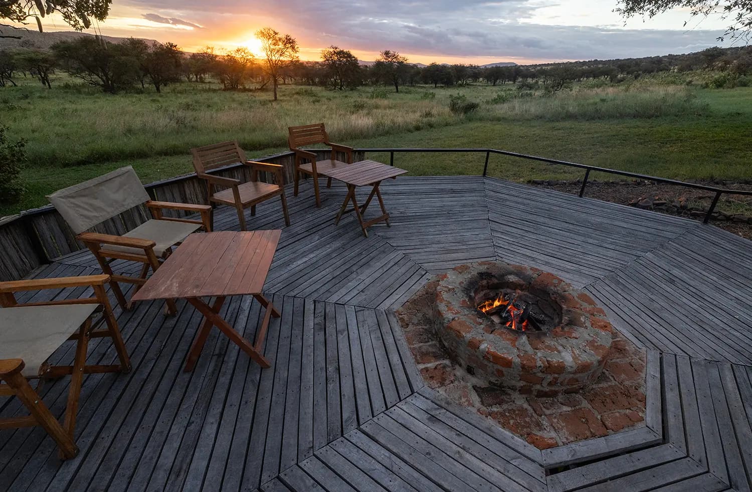 A fire pit on a wooden deck with camp chairs, the sun setting over the plains