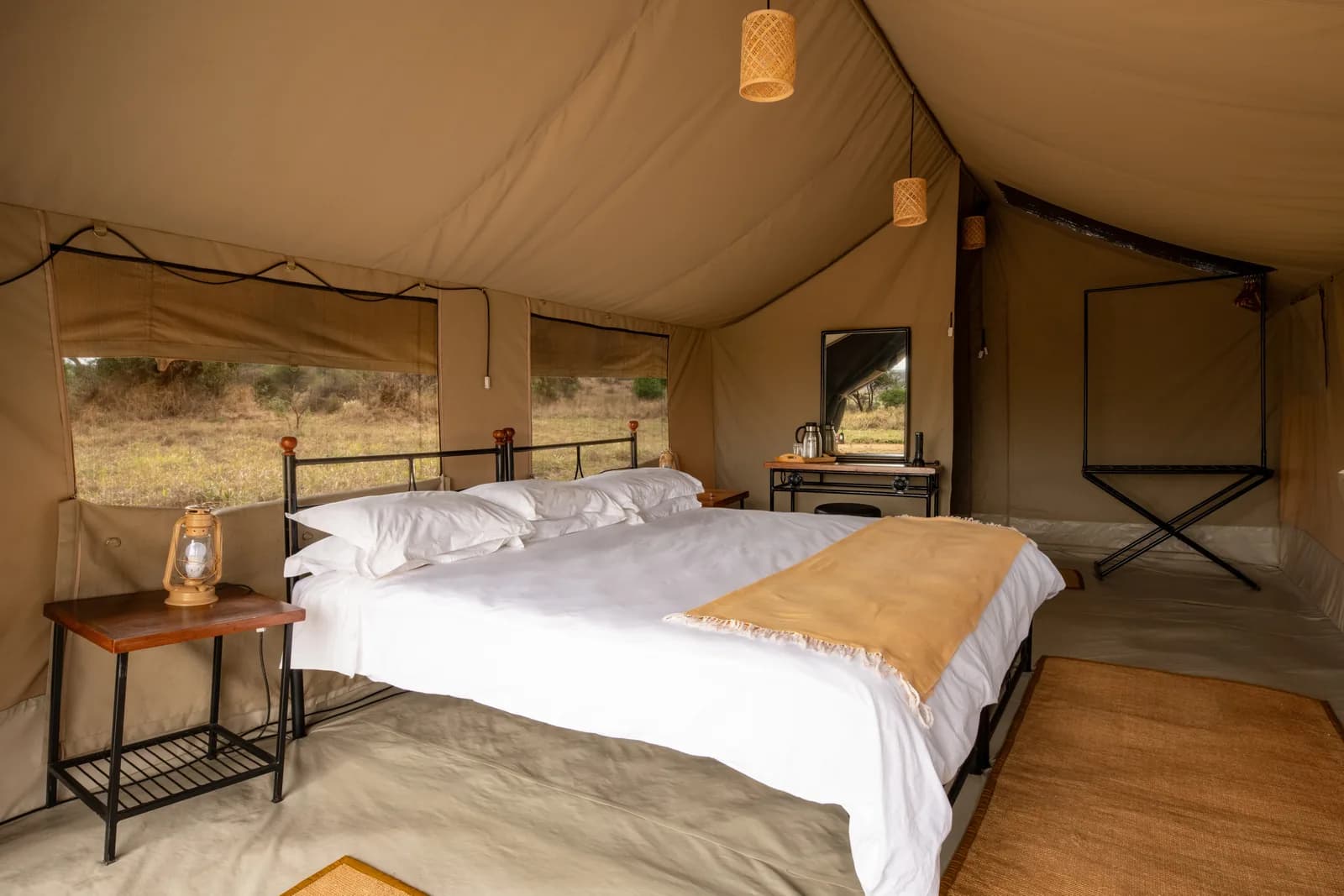 Inside a safari tent: a double bed with a lantern on the bedside table, the bush through the window
