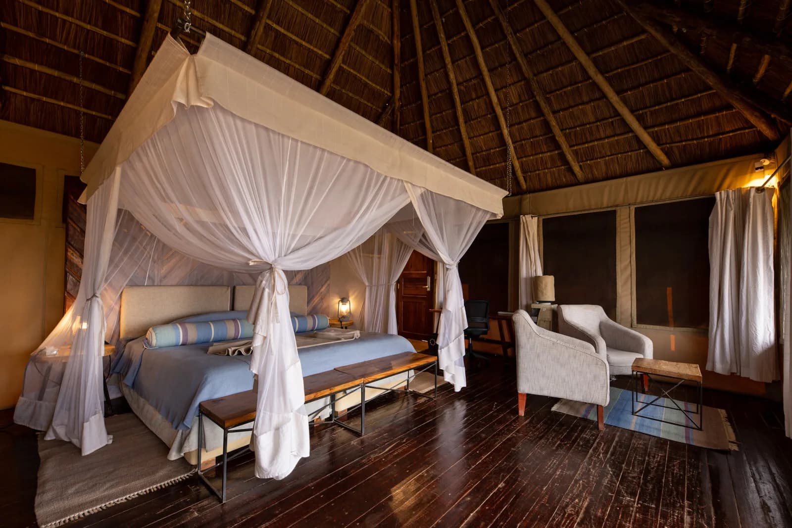 Bedroom with a netted four-poster bed and armchairs under a thatched roof