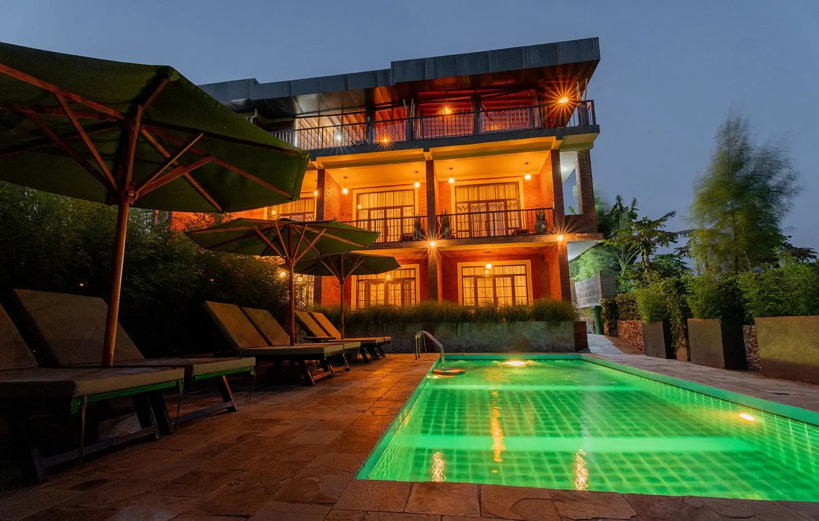 The swimming pool lit green at dusk, with sun loungers and umbrellas in front of the lit-up house