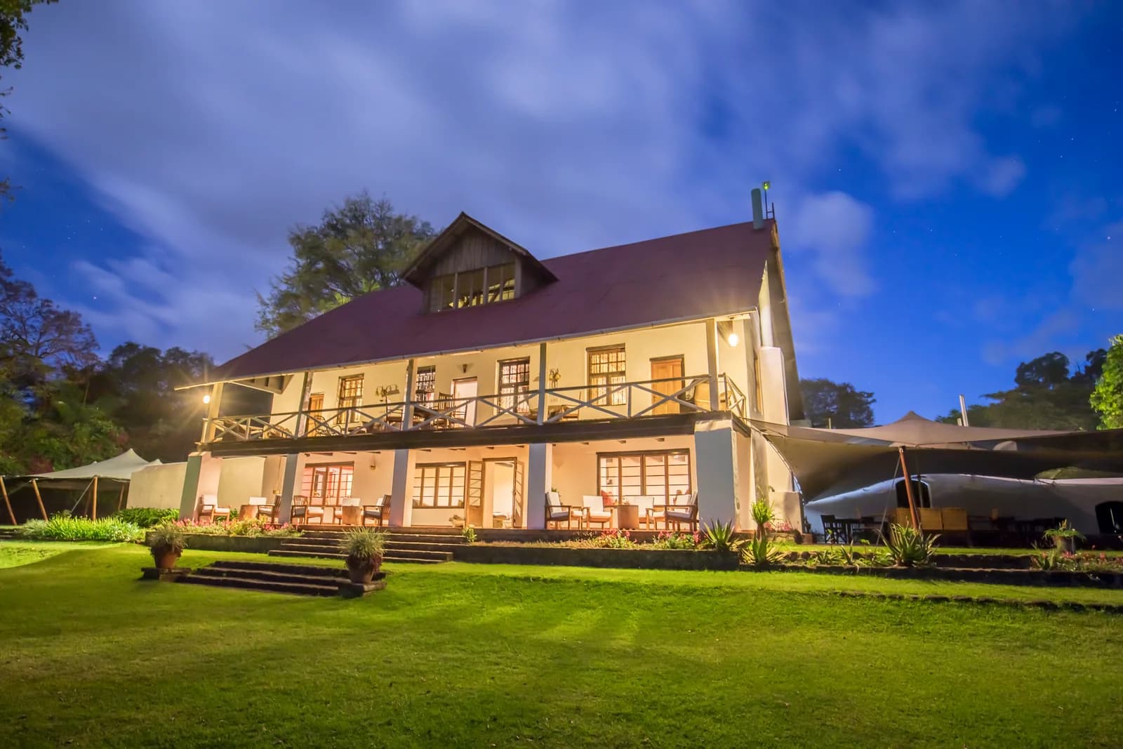 The two-storey main house lit up at dusk, with a veranda above its terrace and a lawn in front
