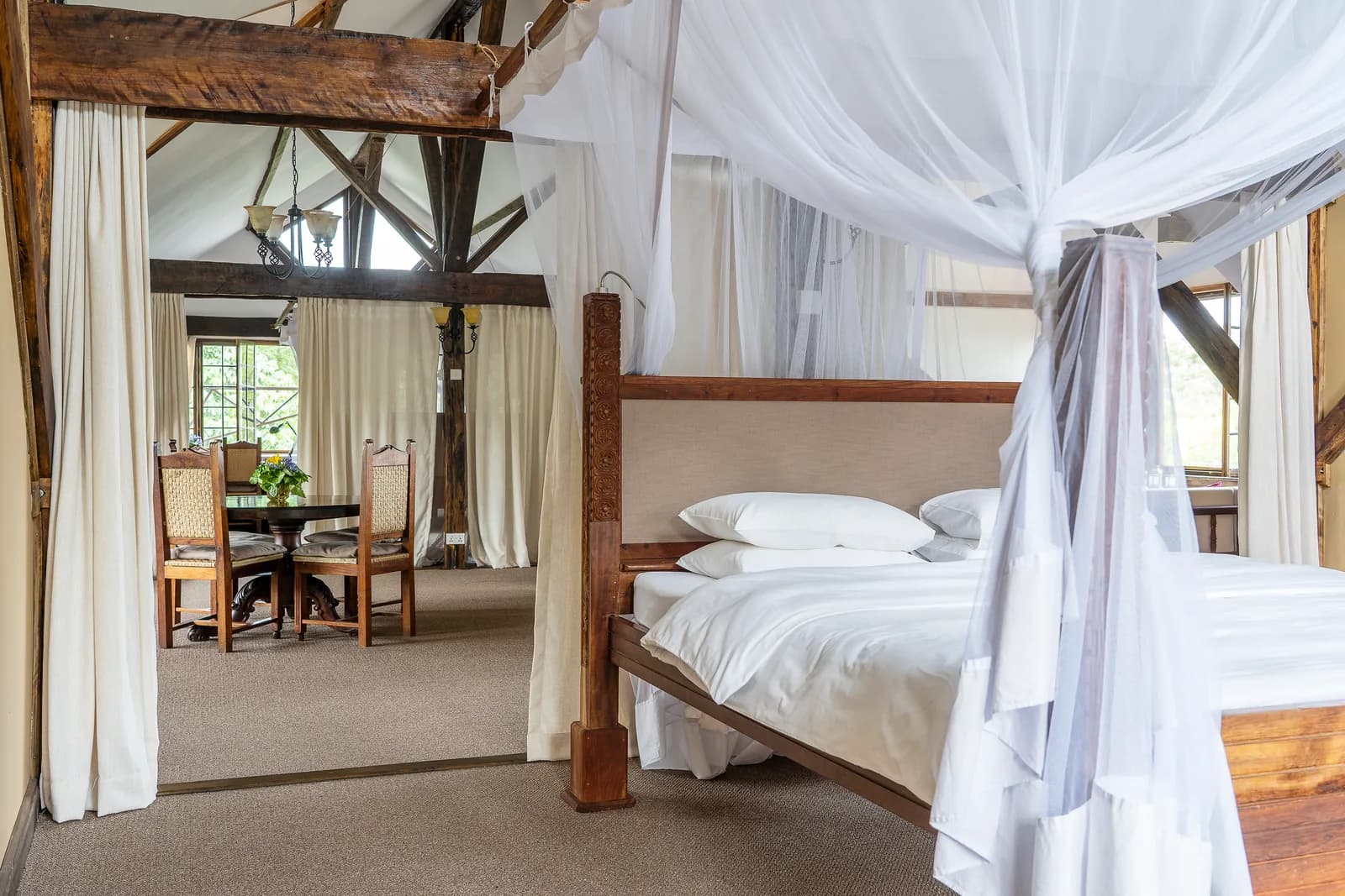 A four-poster bed hung with white netting under exposed roof beams, with a dining table beyond