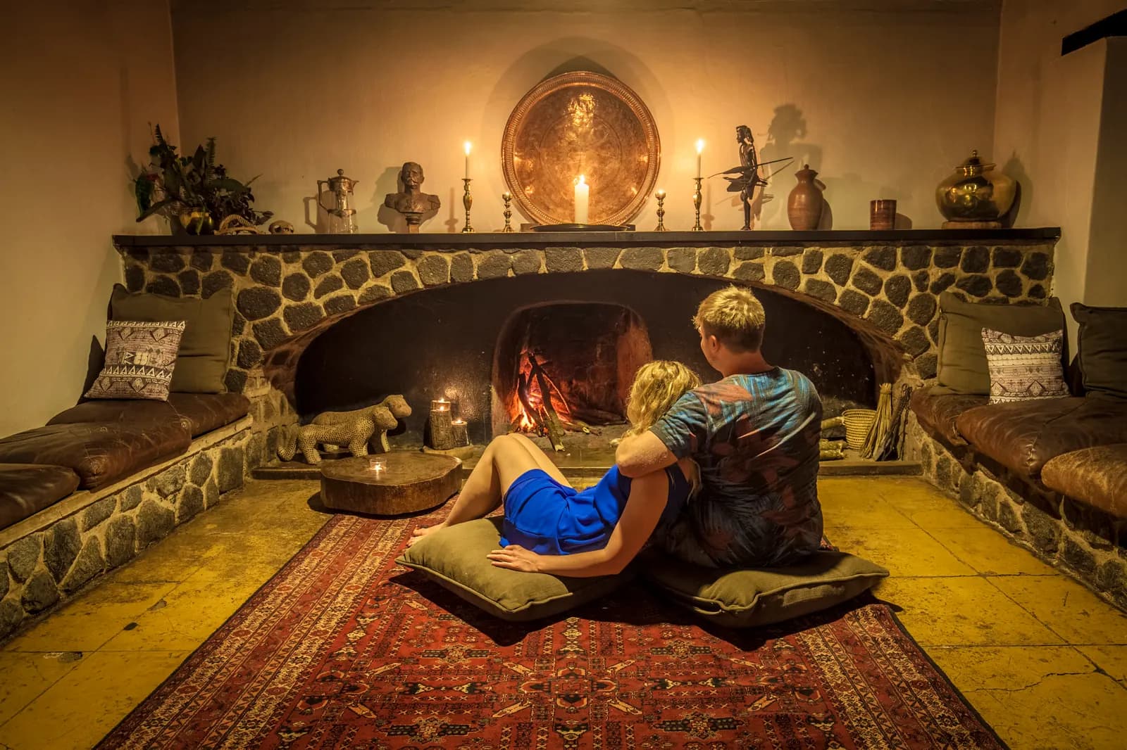 A couple sitting on cushions by a log fire in a stone fireplace, with candles on the mantelpiece