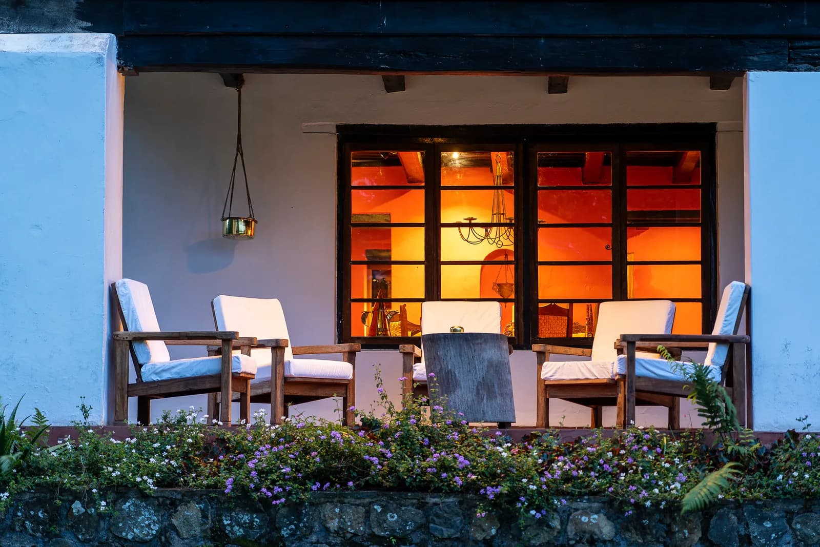 Cushioned chairs on a stone-walled veranda at dusk, in front of a window lit orange from inside